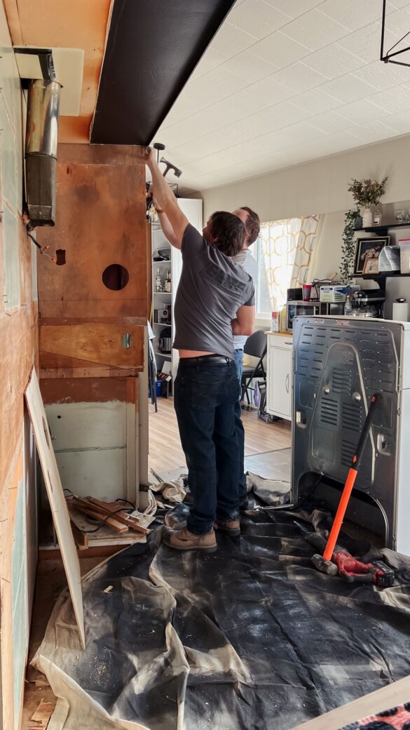 Phase 3 kitchen renovation. Interior Elite Contracting and Joe are working on removing the final cabinet. Most of the cabinets have been removed, exposing the plywood on the walls. There is a drop cloth on the ground covered in wood and various tools. The range is sitting the middle of the kitchen.