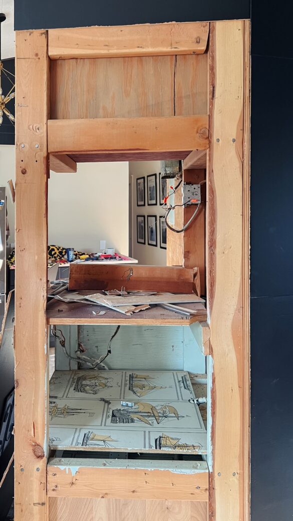 Phase 3 kitchen renovation. A close up showing the skeleton of the cabinets from the 60s. 2 by 4s are exposed, wires are dangling from inside the cabinets. The shelves inside are lined with contact paper covered with sail boats.