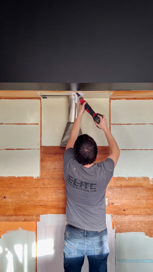 man with brown hair is working with a reciprocating saw to cut a hole in a bulk head. he stands in front of a plywood wall with painted squares, showing where cabinets once were.