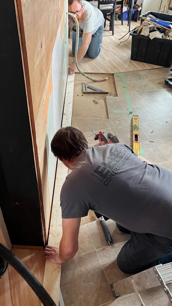 Phase 3 kitchen renovation. Interior Elite Contracting and Joe start framing.  All the cabinets have been removed and the subfloor is now exposed. One person is on one side of the 2 by 4, while the other is at the other end helping measure. 