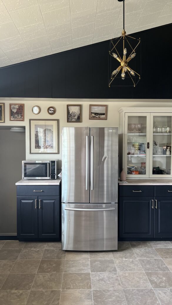 stainless steel fridge sits between two banks of black cabinets. There is a variety of art around the fridge to the left. To the right, there is a beige hutch sitting on the cabinets.