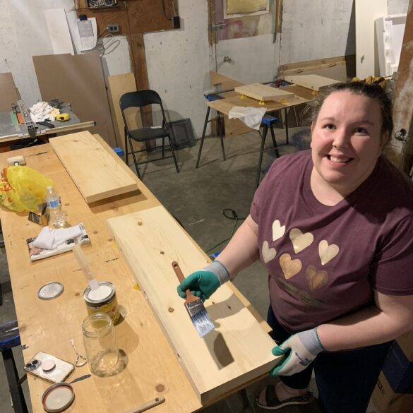 A young 30s caucasian woman with her hair back is in a purple t shirt, wearing gloves and staining wood shelves