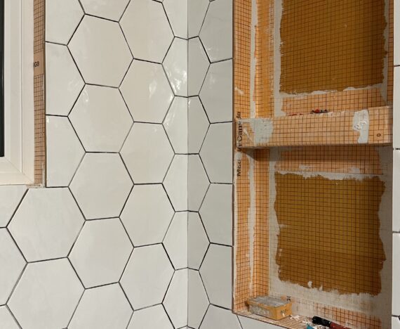 a progress picture of tub surround being redone. The left wall has white hexagon tiles installed, while the wall to its right is only partially tiled with two niches for space for shampoo and conditioner bottles