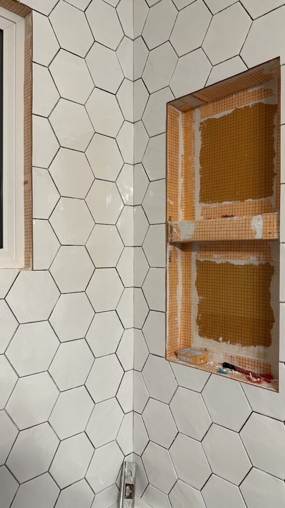 a progress picture of tub surround being redone. The left wall has white hexagon tiles installed, while the wall to its right is only partially tiled with two niches for space for shampoo and conditioner bottles