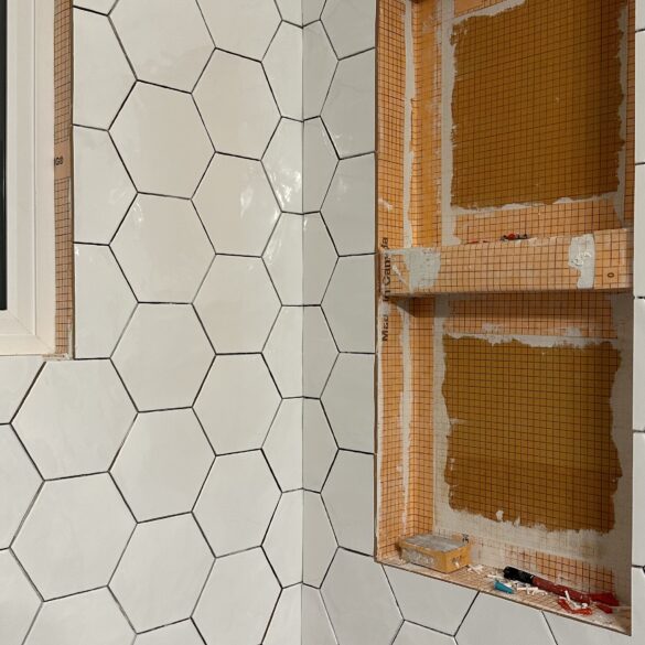a progress picture of tub surround being redone. The left wall has white hexagon tiles installed, while the wall to its right is only partially tiled with two niches for space for shampoo and conditioner bottles