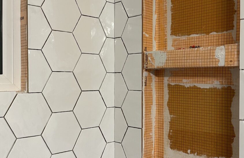 a progress picture of tub surround being redone. The left wall has white hexagon tiles installed, while the wall to its right is only partially tiled with two niches for space for shampoo and conditioner bottles